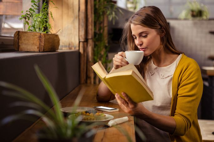A woman sits at a cafe and reads with a warm drink. reading, woman reading, solo traveler, book, cafe